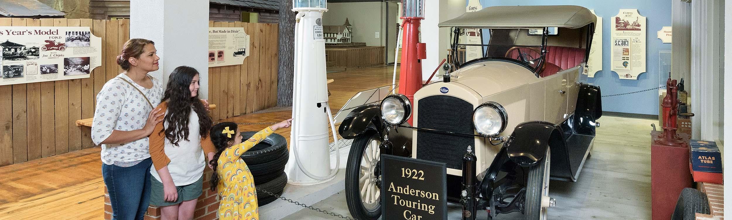 Mom and 2 daughters stand look at a white, early 20th century vintage car on display at the SC State Museum