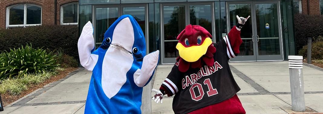 Person in blue shark mascot costume stands next person in Gamecock mascot uniform in front of a building entrance that reads SCSM