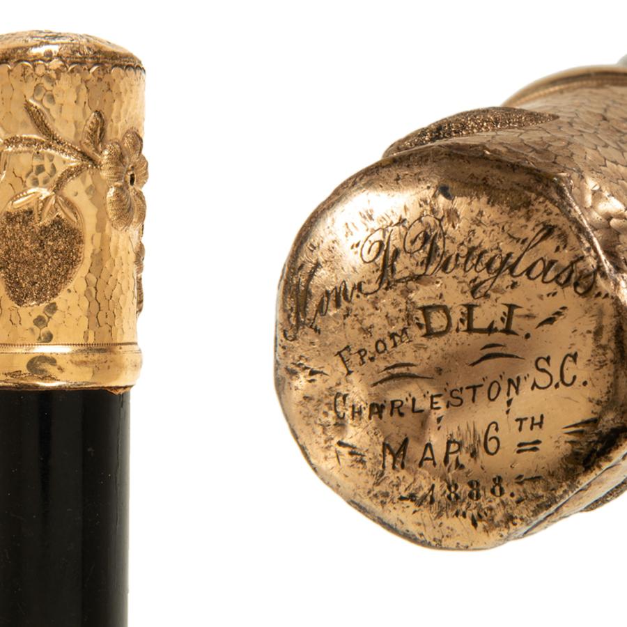 Showing the top of a walking stick. It is gold with fruit on the side and text on the top.