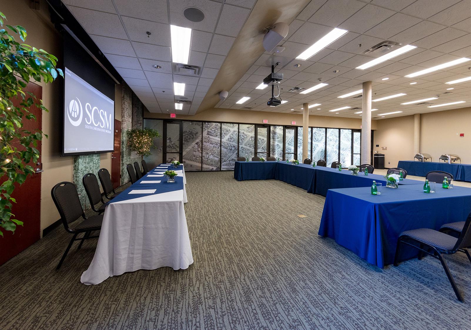 Large room with tables set up in u shape facing another line of tables and projector screen 