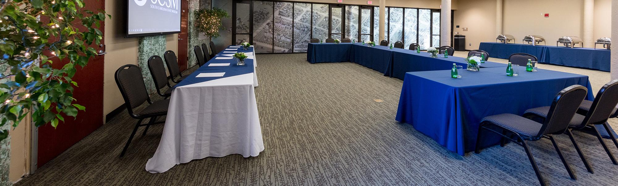 Large room with tables set up in u shape facing another line of tables and projector screen 