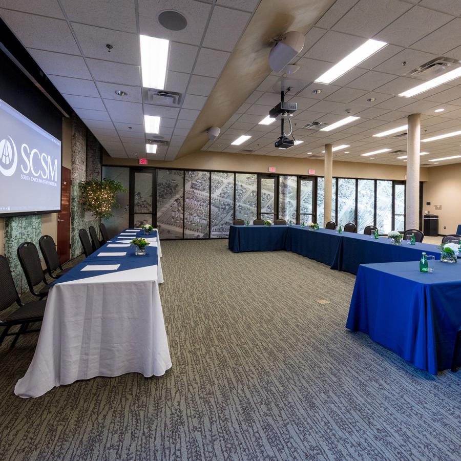 Large room with tables set up in u shape facing another line of tables and projector screen 