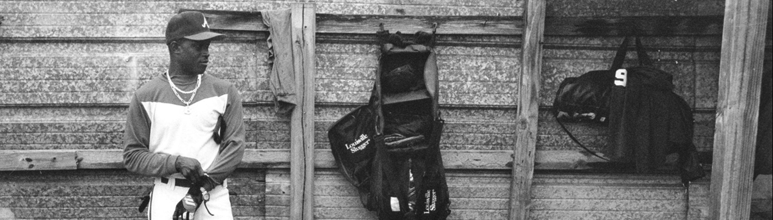 African-American baseball player stands against a wooden wall as he pulls on his gloves to prepare for his time at bat.