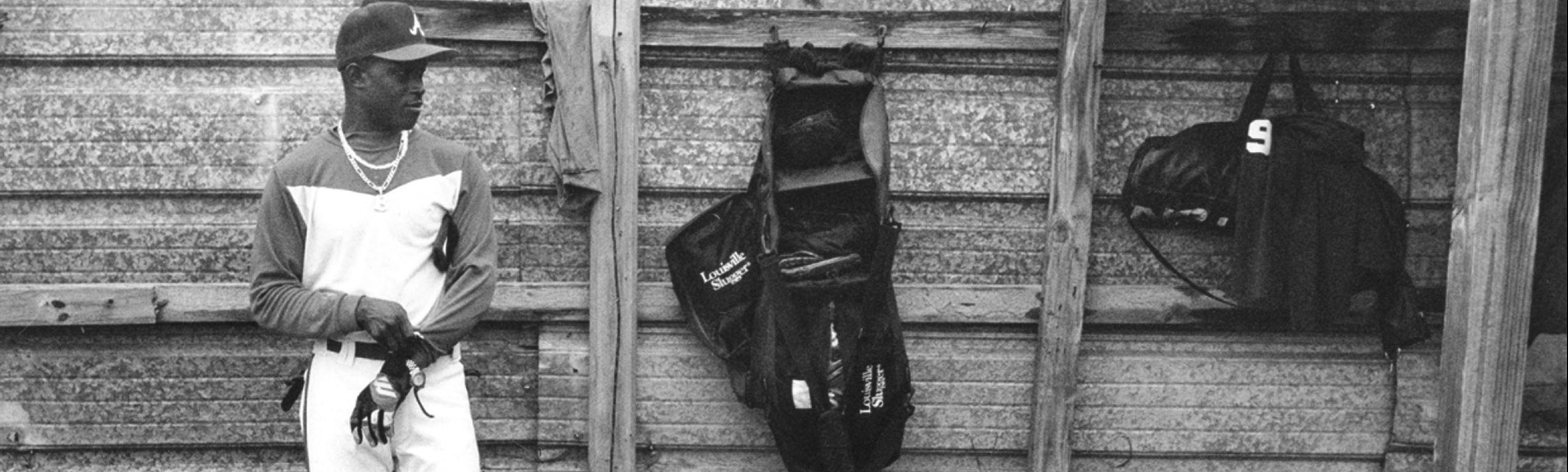 African-American baseball player stands against a wooden wall as he pulls on his gloves to prepare for his time at bat.