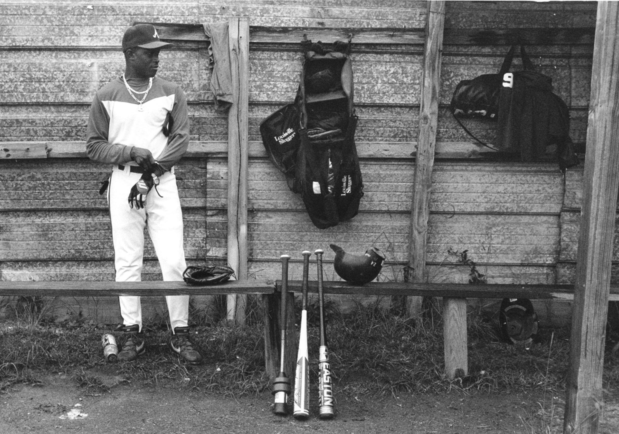 African-American baseball player stands against a wooden wall as he pulls on his gloves to prepare for his time at bat.