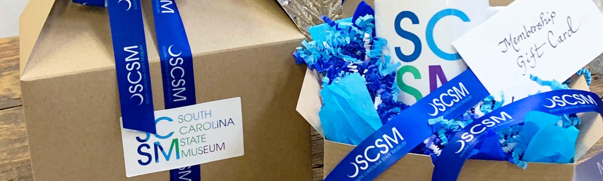 Two brown gift boxes on display with one partly open to show an SCSM mug inside with blue SCSM branded ribbon entwined