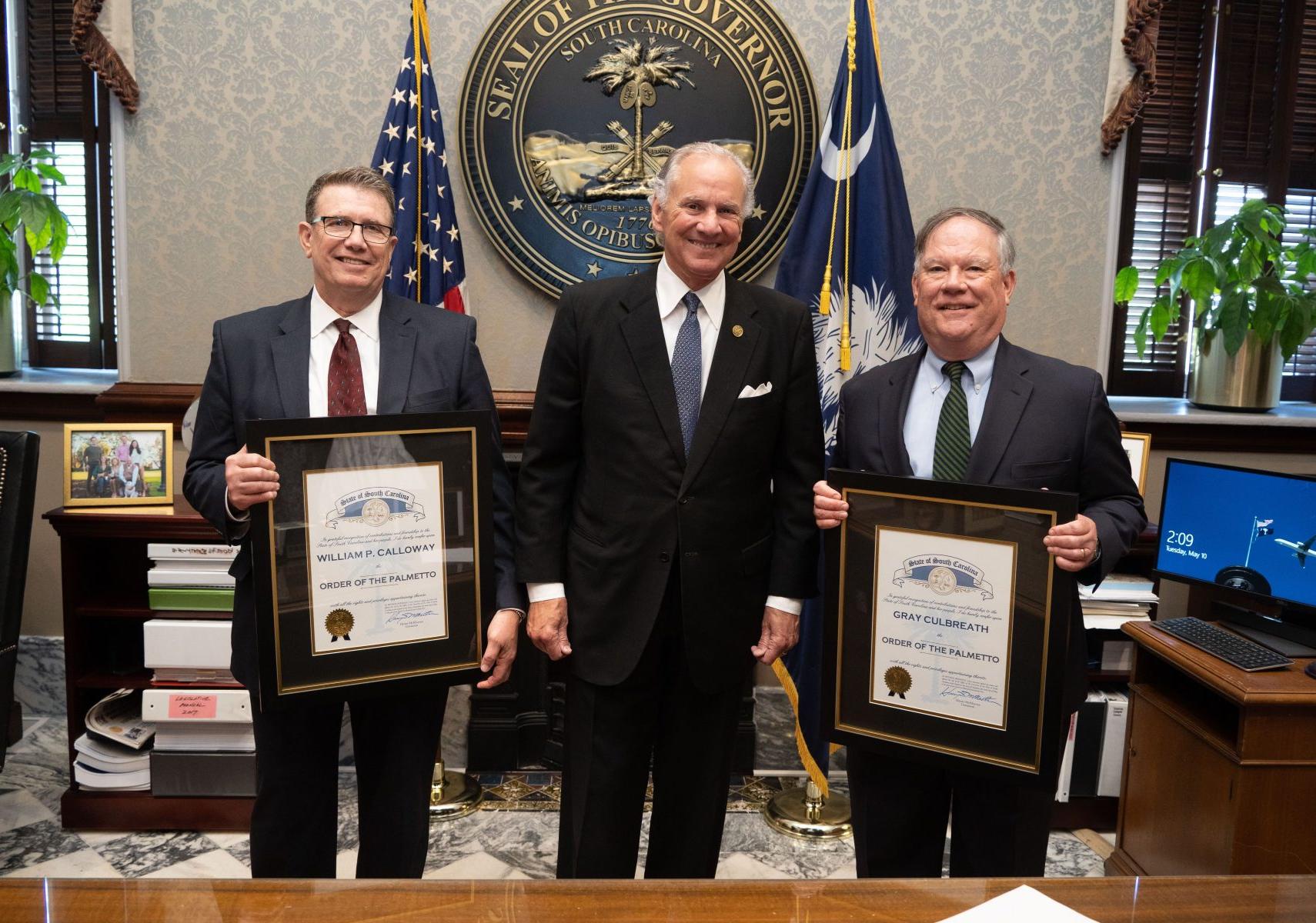 Three men standing, two holding awards