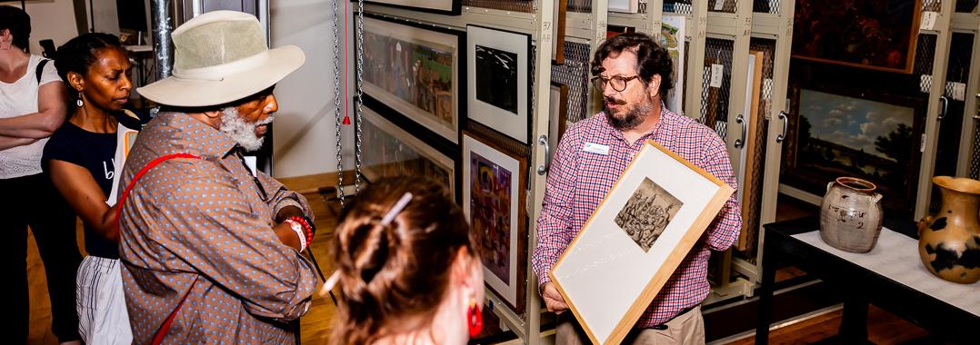 Man with beard hold up a framed drawing so it can be seen by the group of 4 people facing him. Racks of paintings are visible in the background.