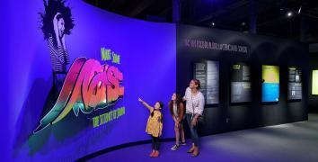 A Mother with two daughters look at the exhibit entrance which features a large sign with the exhibit title done in a graffiti art style.