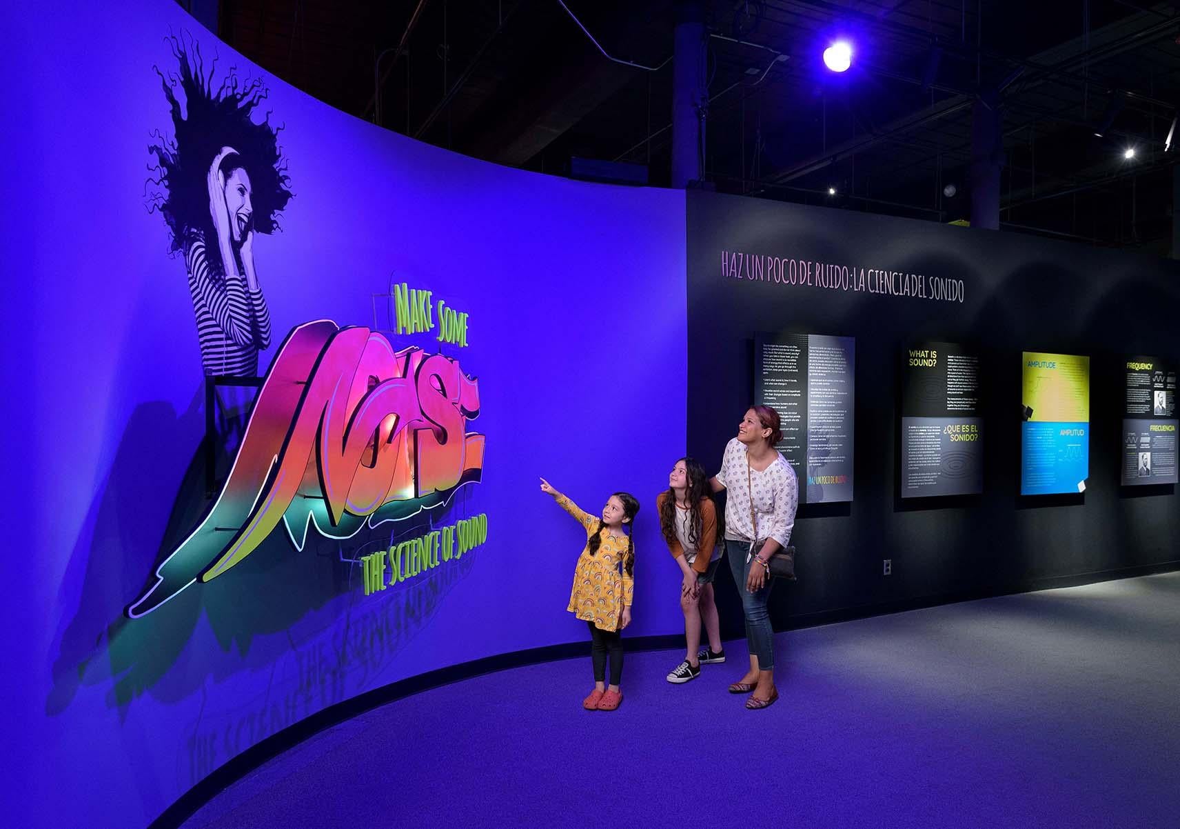 A Mother with two daughters look at the exhibit entrance which features a large sign with the exhibit title done in a graffiti art style.