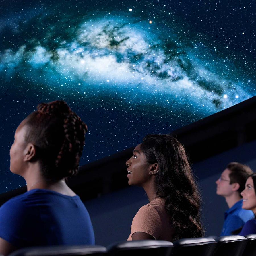 Close up image of four people shown from a back/side profile as they look up to watch a projection of the Milky Way in a planetarium dome