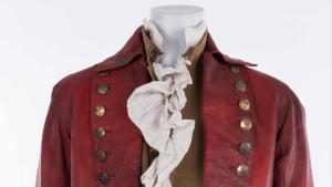 Mannequin on white background wearing red knee length coat, a tan vest and frilly white shirt