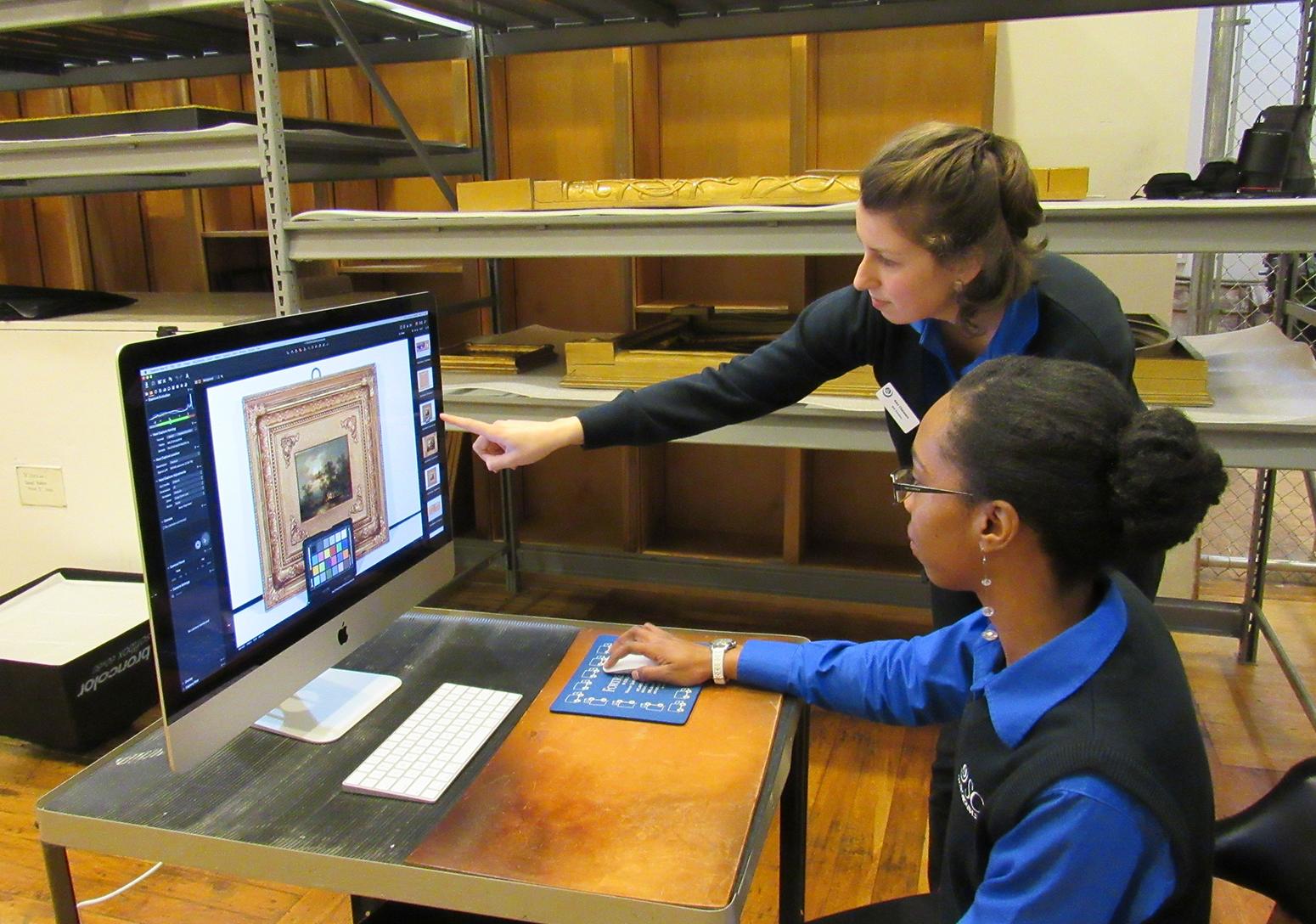 Two women, one standing and one seated wearing blue sweaters point at a computer screen that has a photograph of a painting on it.