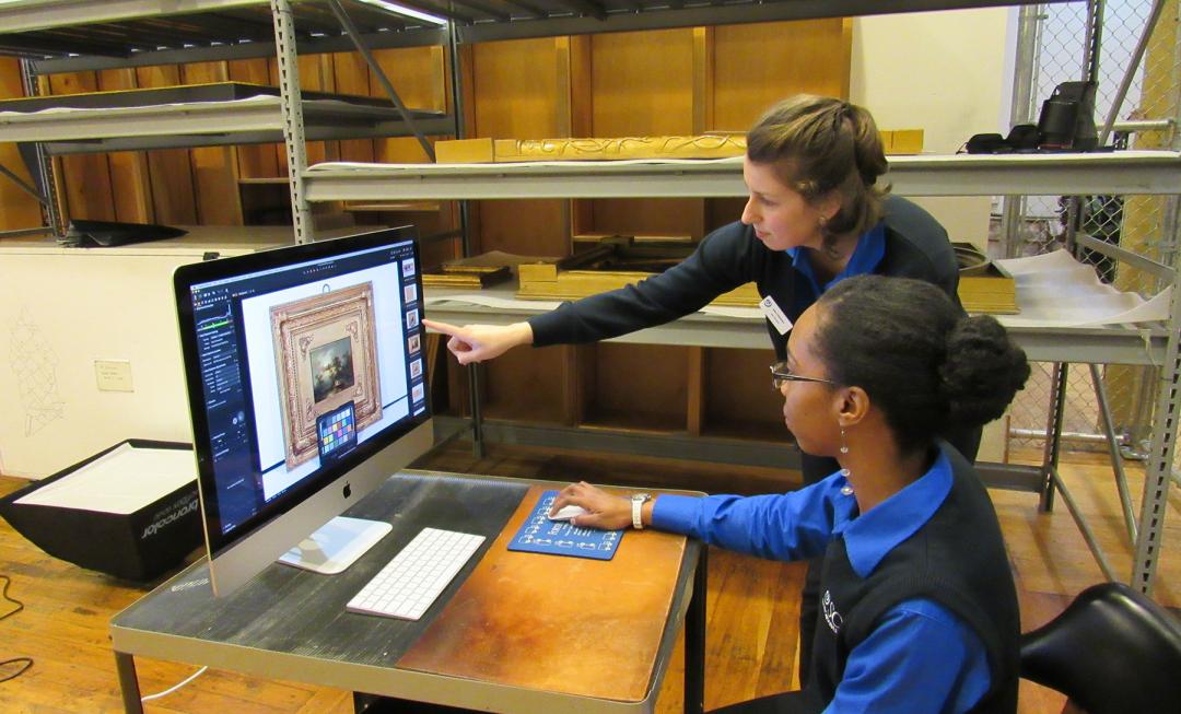 Two women, one standing and one seated wearing blue sweaters point at a computer screen that has a photograph of a painting on it.