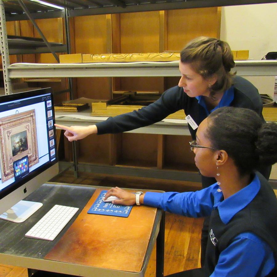 Two women, one standing and one seated wearing blue sweaters point at a computer screen that has a photograph of a painting on it.