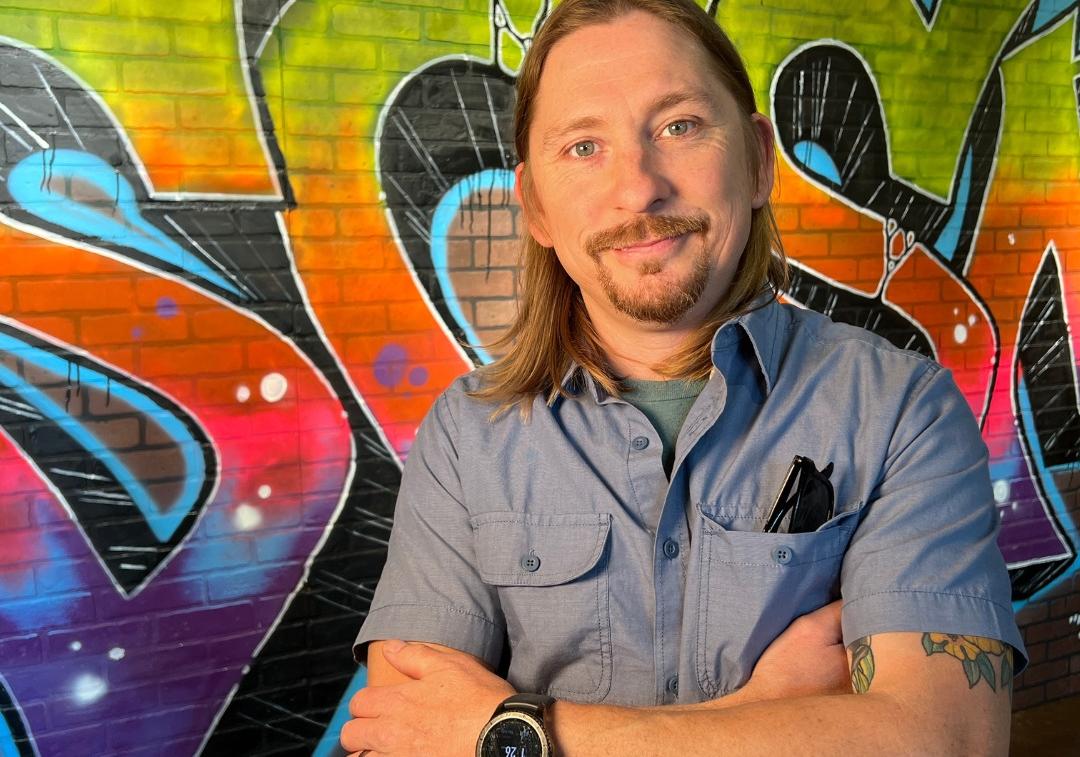 Man with shoulder length hair and mustache stands with arms crossed smiling at camera with wall in background that has a spray painted SCSM on it