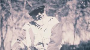 Black and white photo of man seated on tree trunk with large beard and wearing sunglasses and a flat cap sunglasses and