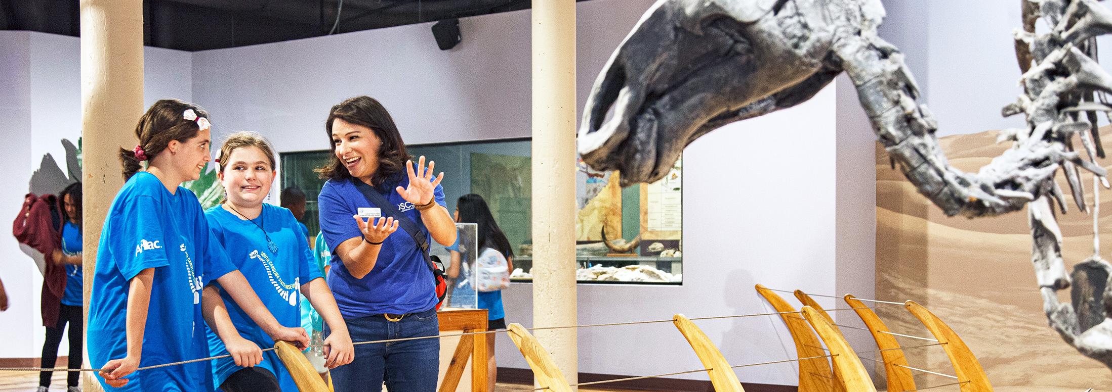 2 young girls with bright blue t-shirts stand next a women in a matching t-shirt who is pointing towards a dinosaur skeleton on display