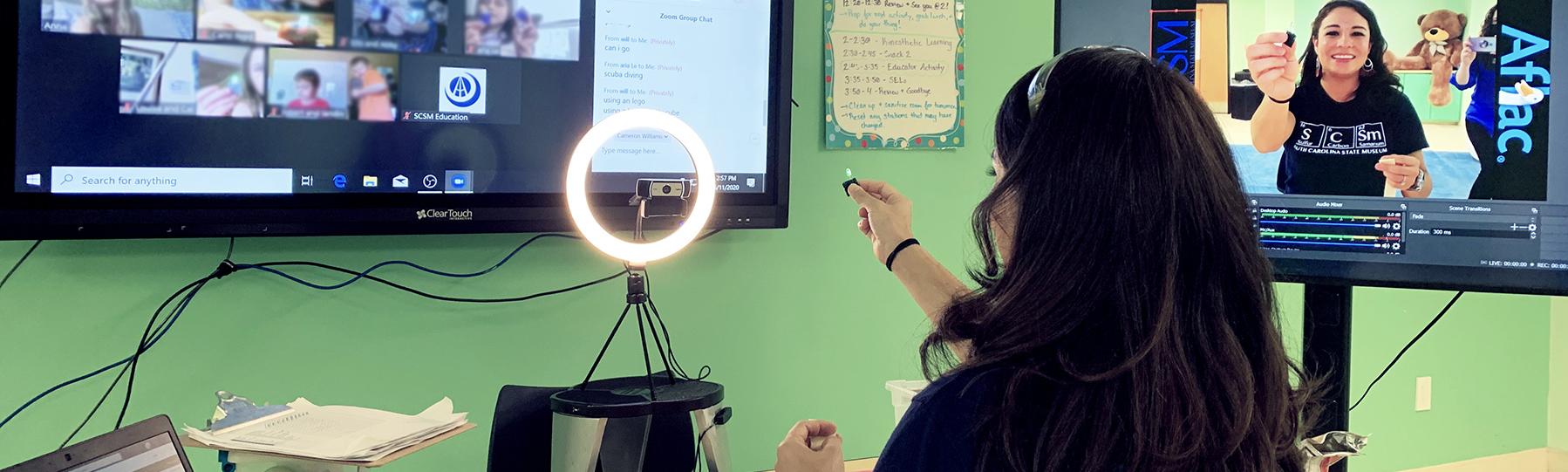Museum educator with dark hair sits before two large digital screens displaying live video of students watching her teach a lesson.
