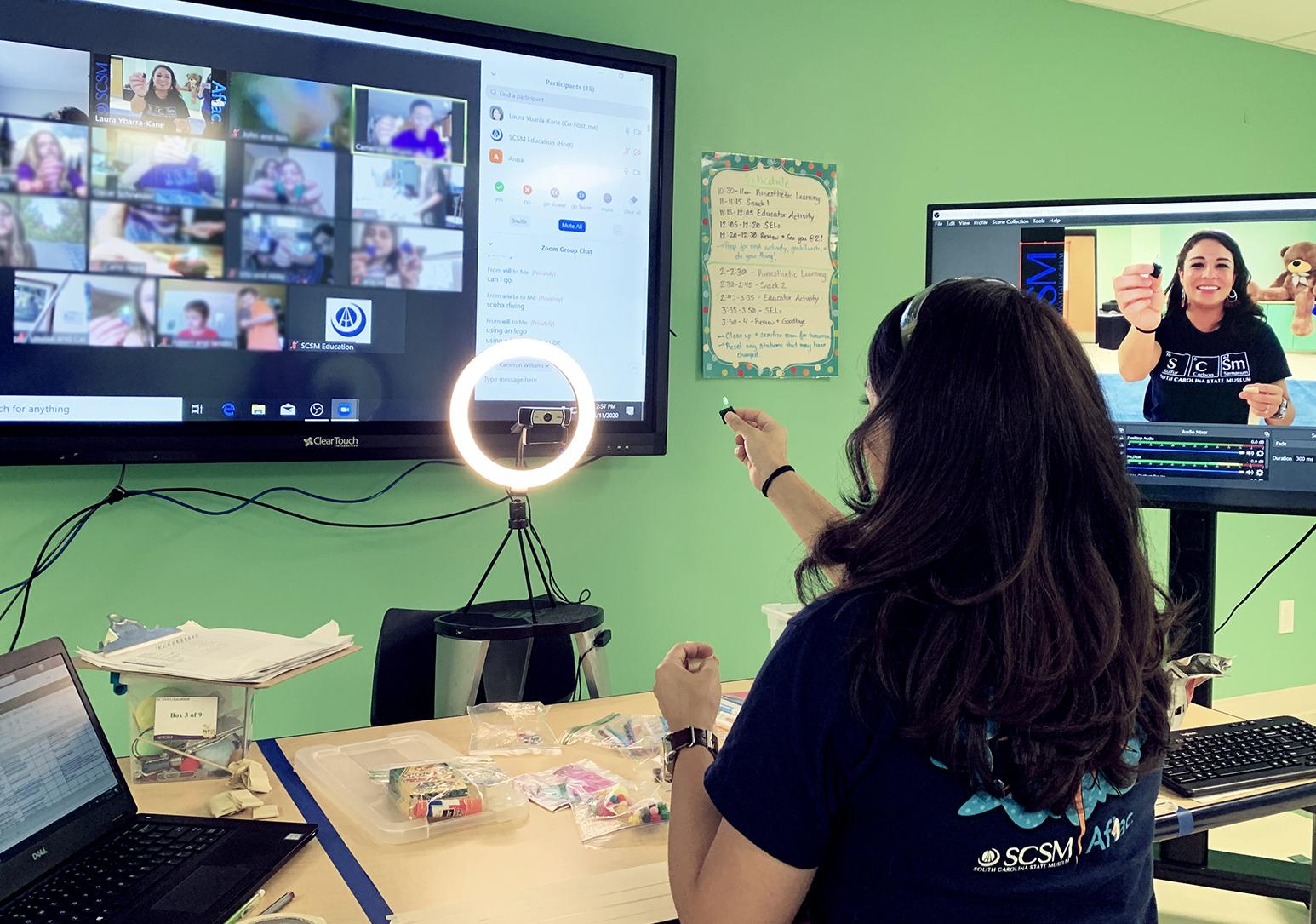 Museum educator with dark hair sits before two large digital screens displaying live video of students watching her teach a lesson.