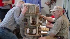 Two men holding a concrete replica of a classical tower as they try to reposition it