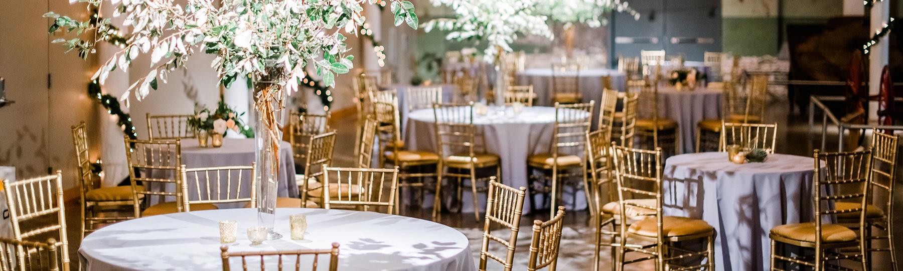 Large space with string lights suspended from ceiling and round tables with silver table cloths, gold chairs and large flower center pieces.