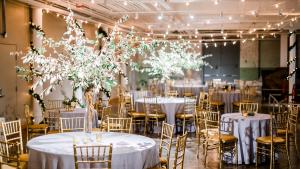 Large space with string lights suspended from ceiling and round tables with silver table cloths, gold chairs and large flower center pieces.