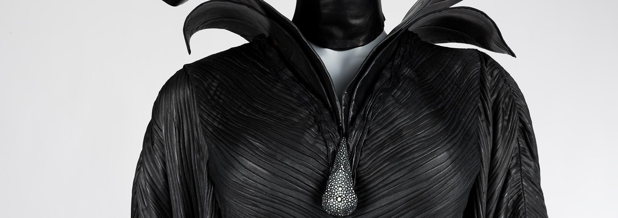 Close up of a black dress with textured fabric, a large tear drop in dark silver gems decorate the neckline with a collar that stands up and out over the shoulders like wings
