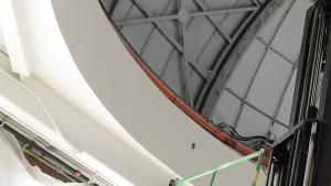 Large white telescope tube suspended from life as man on ladder guides it into place on a large base. Other staff are nearby on a bucket lift and on a forklift.