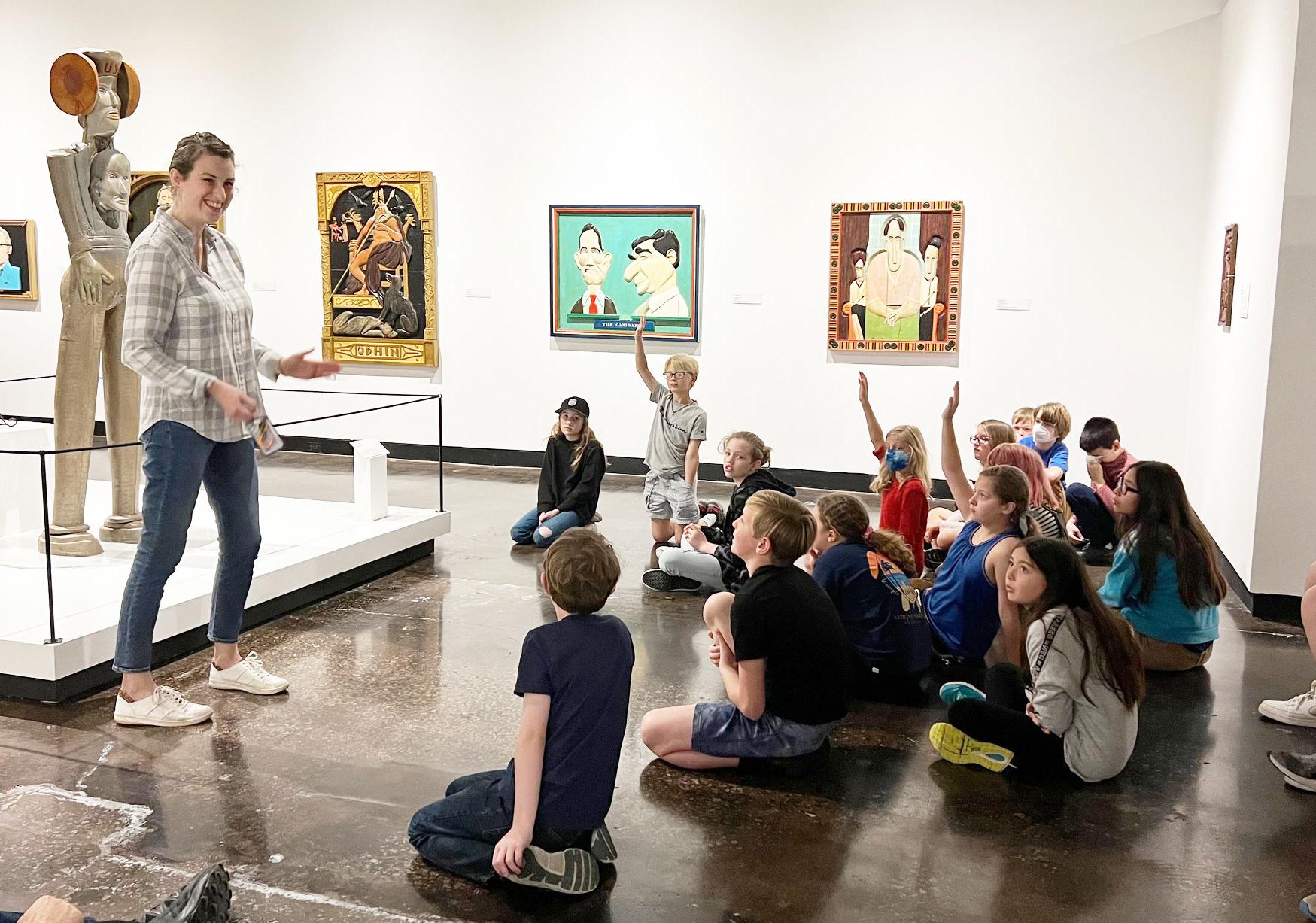 Curator of Art stands in from of a group of children, some raising their hands, seated on the floor in the Lipscomb Art Gallery. 