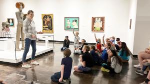 Curator of Art stands in from of a group of children, some raising their hands, seated on the floor in the Lipscomb Art Gallery.
