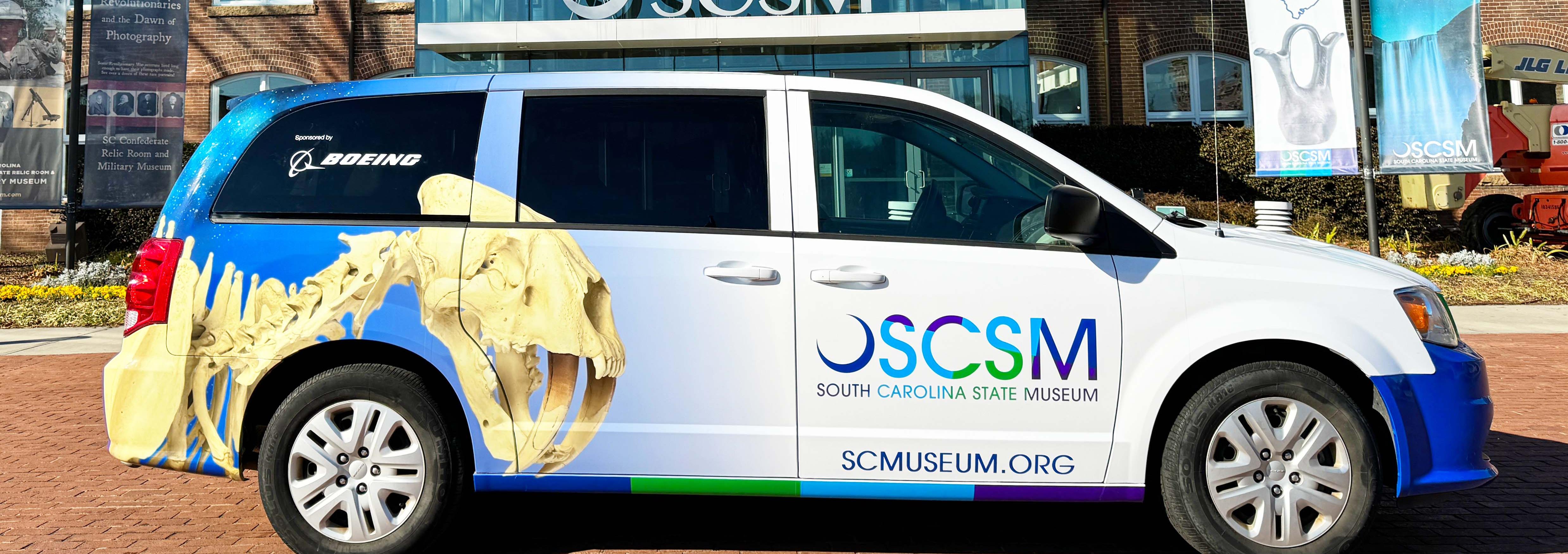 Mini van parked in front of the large glass museum entrance. Van features the image of a sabercat fossil along its side.