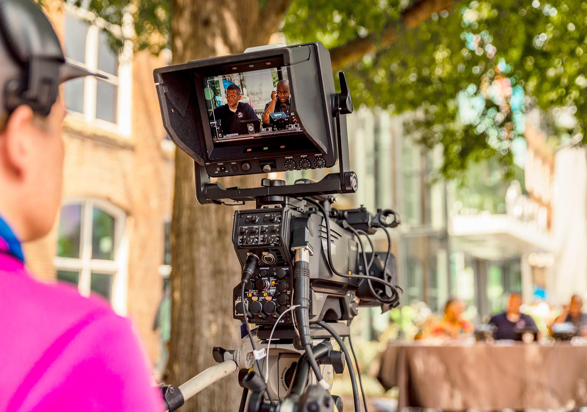 Man wearing hat and headphones with back facing the viewer operates a large television camera.