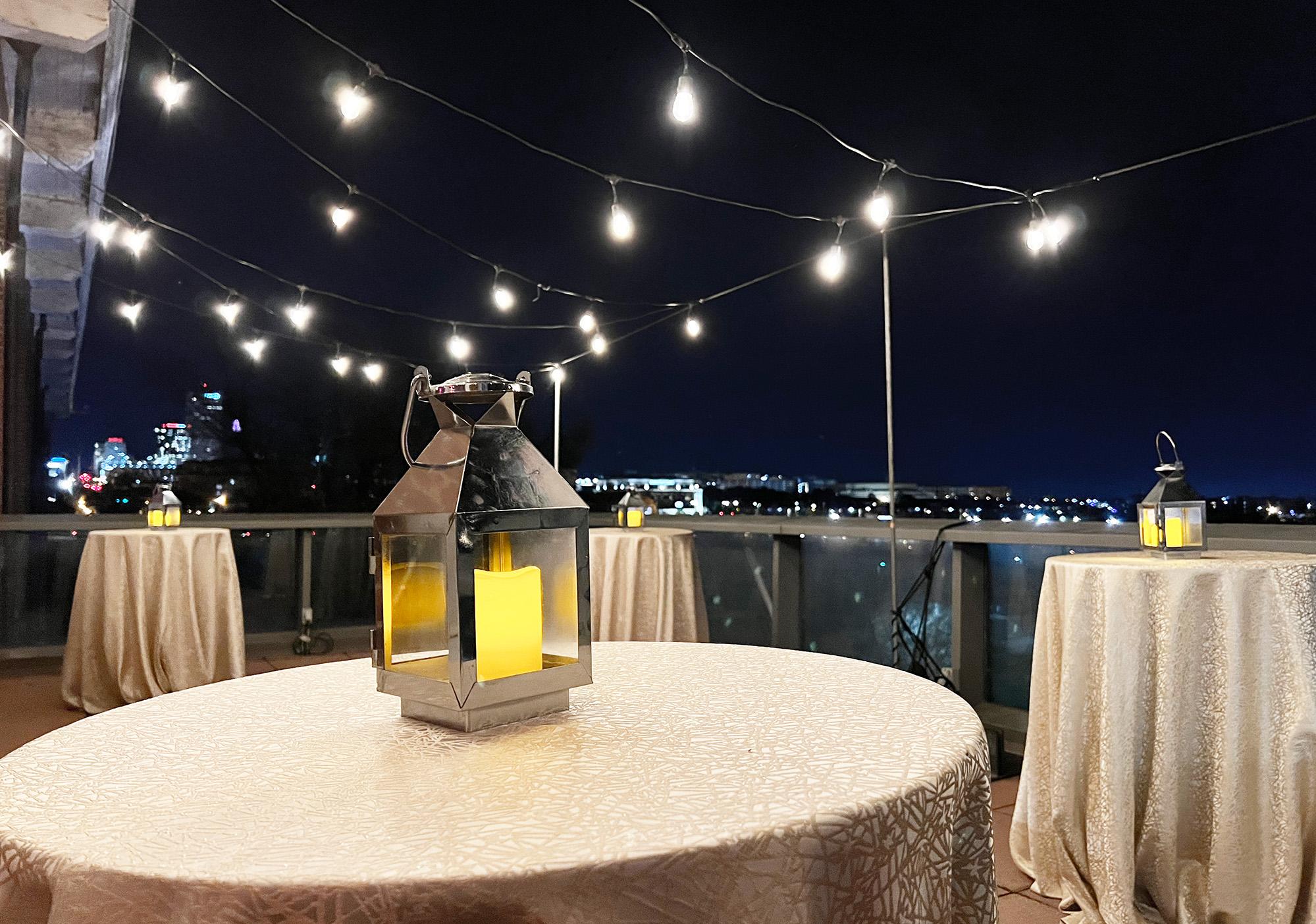 Terrace at night with skyline visible in background and string lights set up above three formally set tables