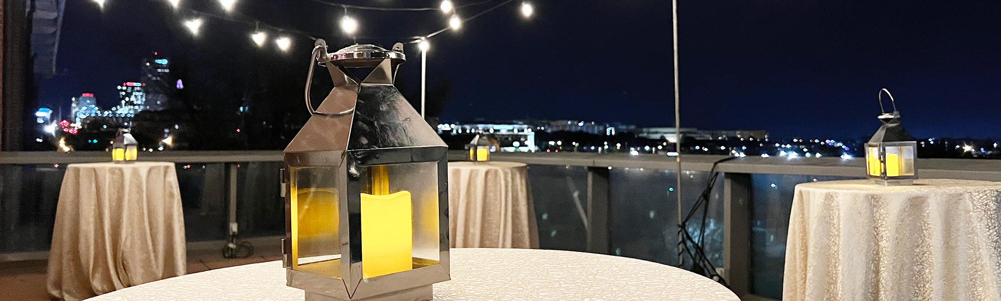 Terrace at night with skyline visible in background and string lights set up above three formally set tables