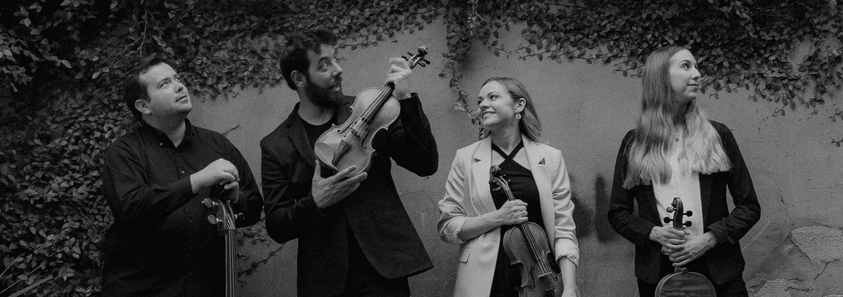 Black and white with 2 women and 2 men. One man holds a cello and the other all hold violins. They are standing in front of an ivy covered wall.