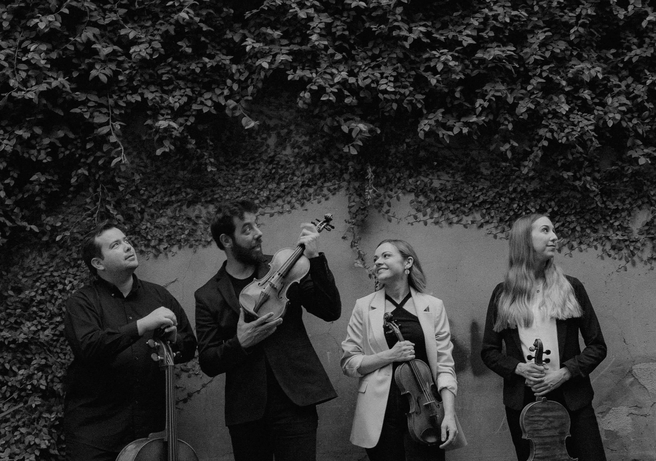 Two men in dark suits and two women in dark clothes with one in a cream colored blazer and the other in a black blazer. They are all standing on a street with a brick building behind them. One man holds a cello and the others all hold violins.