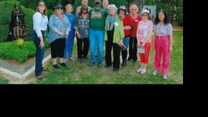 Group of 11 older people in casual clothing stand pose for a photo with an older black man who is in the center of the group. Green hedges and plants are visible in the background.