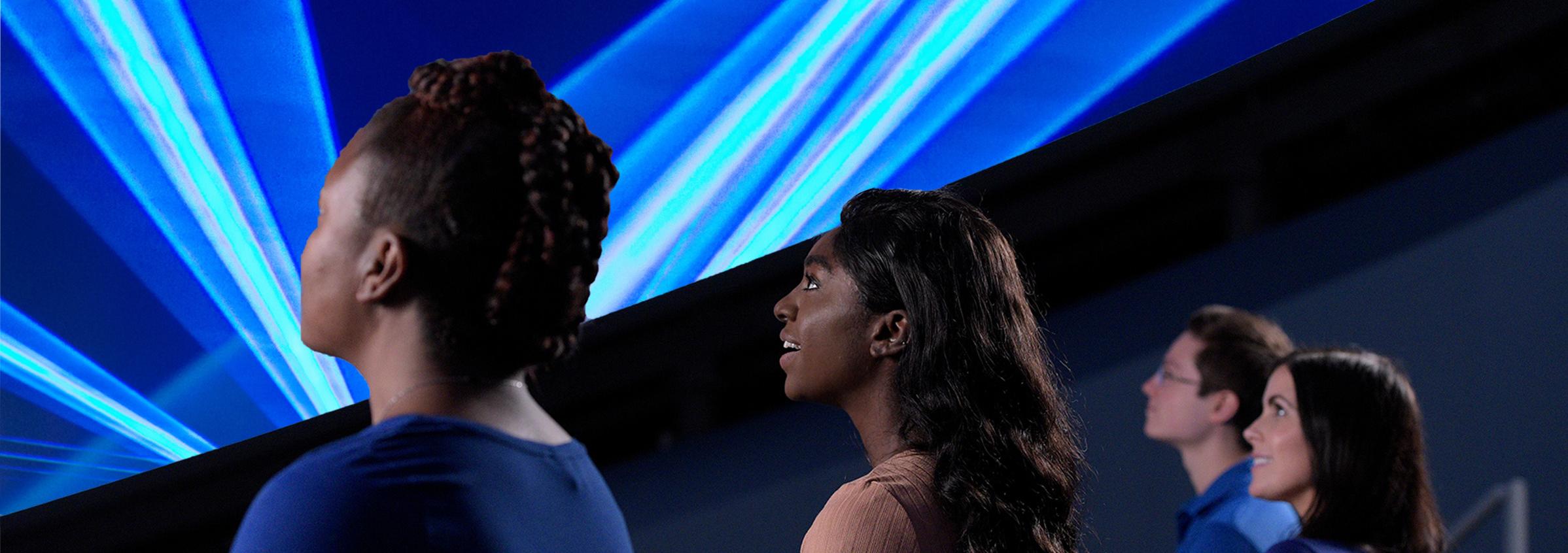 Guests in audience look up and a large, domed screen with blue lasers across it