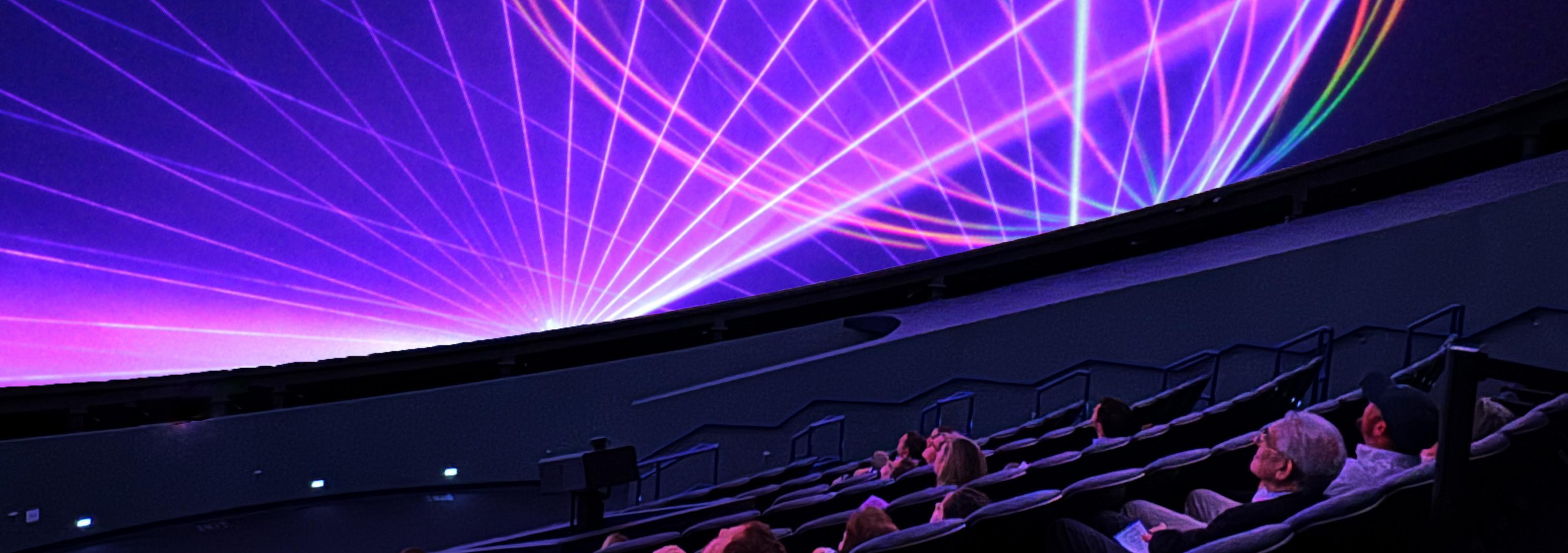 Seated audience looking up a domed screen with pink and purple lasers dancing across it