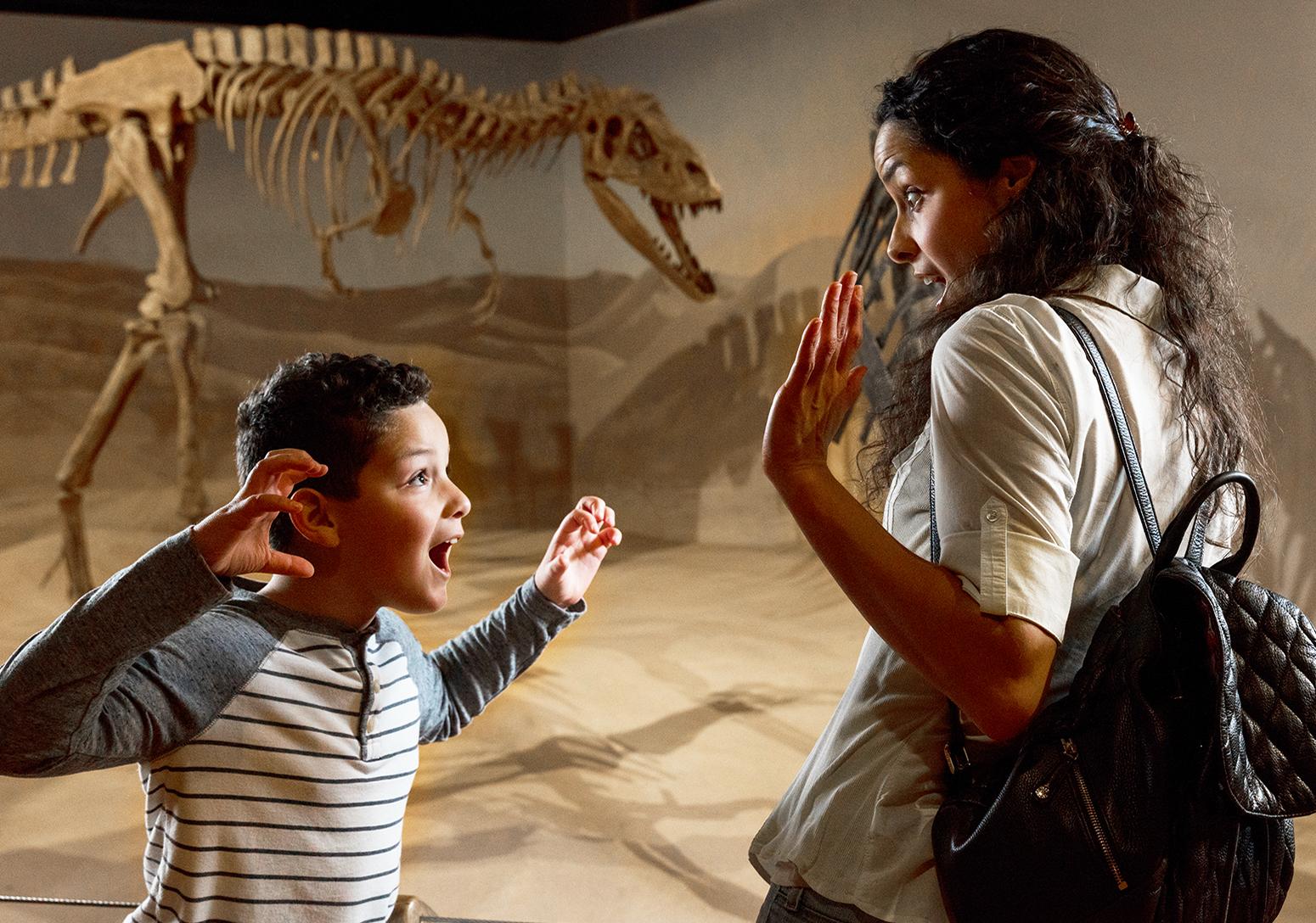 Woman and young boy stand in front of dinosaur skeleton as the boy makes a 'Rawr' face with hands up like dinosaur claws in the direction of the woman