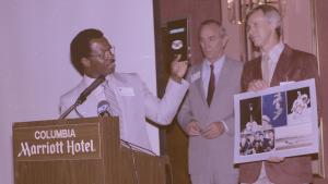 Faded color photograph shows a black man with a mustache in gray suit holding up a dark blue notebook with a NASA logo on the front. He is standing behind a wood panel podium that reads &amp;quot;Columbia Marriot&amp;quot; in white on the front. There are two older men standing just behind the speaker at the podium, one is hold a placard with images of space and astronauts on it.