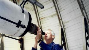 Man in blue shirt looks through eye piece of large white telescope