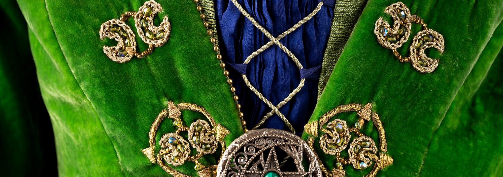 Mannequin on a white bClose up image of a green velvet dress with gold embroidered detailing and a green cape.