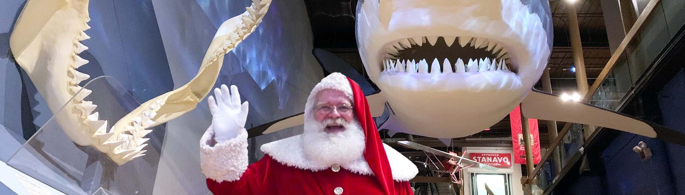 Man in red suit and cap with white trim and fluffy white beard stands and waves with a large replica shark visible behind him