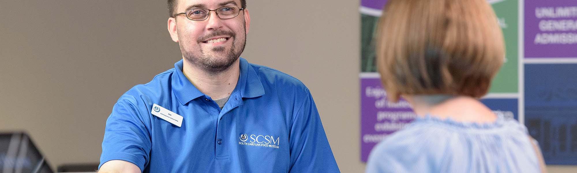 Man with short beard wears blue shirt with museum logo and hands tickets to woman facing away from camera