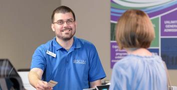 Man with short beard wears blue shirt with museum logo and hands tickets to woman facing away from camera