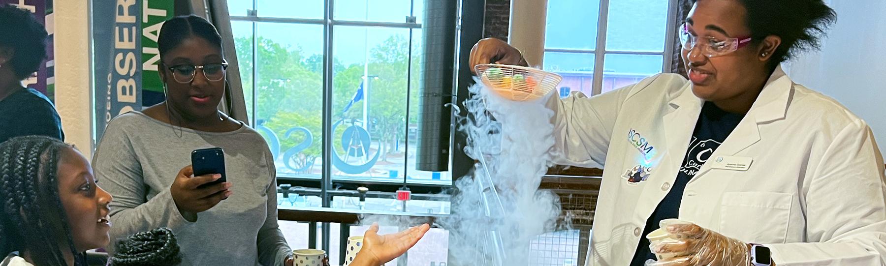 Woman with lab coat holds ladle above a metal bowl with fog overflowing the blow while two young girls watch and one reaches out towards the fog