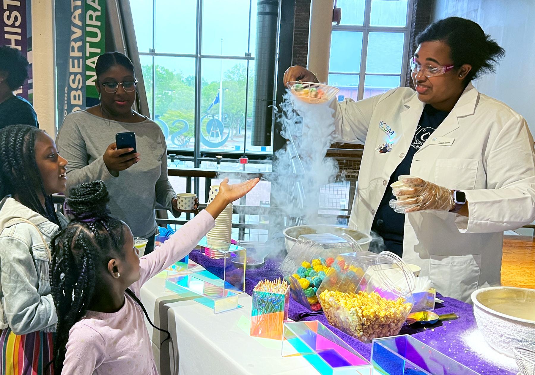 Woman with lab coat holds ladle above a metal bowl with fog overflowing the blow while two young girls watch and one reaches out towards the fog