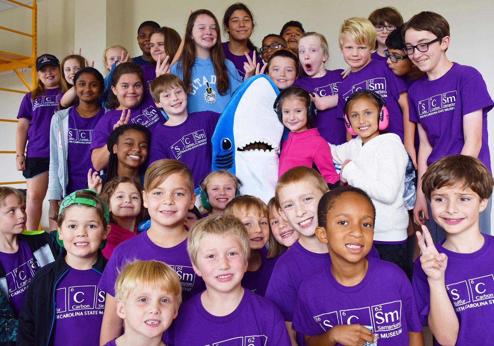 Large group of kids aged 6-12 wearing purple SCSM camp shirts posing with an educator in a shark mascot costume.